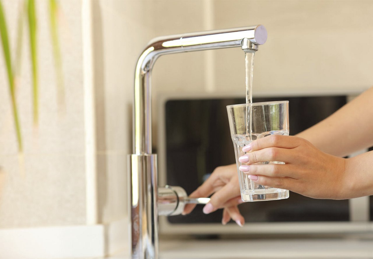 A person filling a glass with water at a kitchen faucet, holding the glass under the running water while their hand turns the handle.