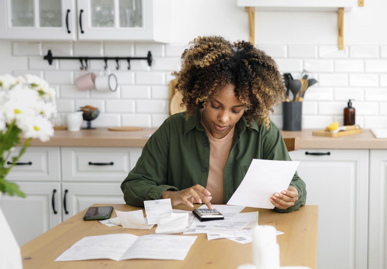 A woman sitting at a kitchen table in her apartment, surrounded by papers and typing in a calculator, with stylish kitchen decor and appliances in the background.