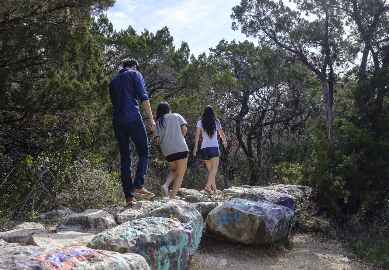 A group of young adults hiking together in a park in Austin, Texas, surrounded by colorful artwork on the rocks and lush trees, enjoying their outdoor adventure.