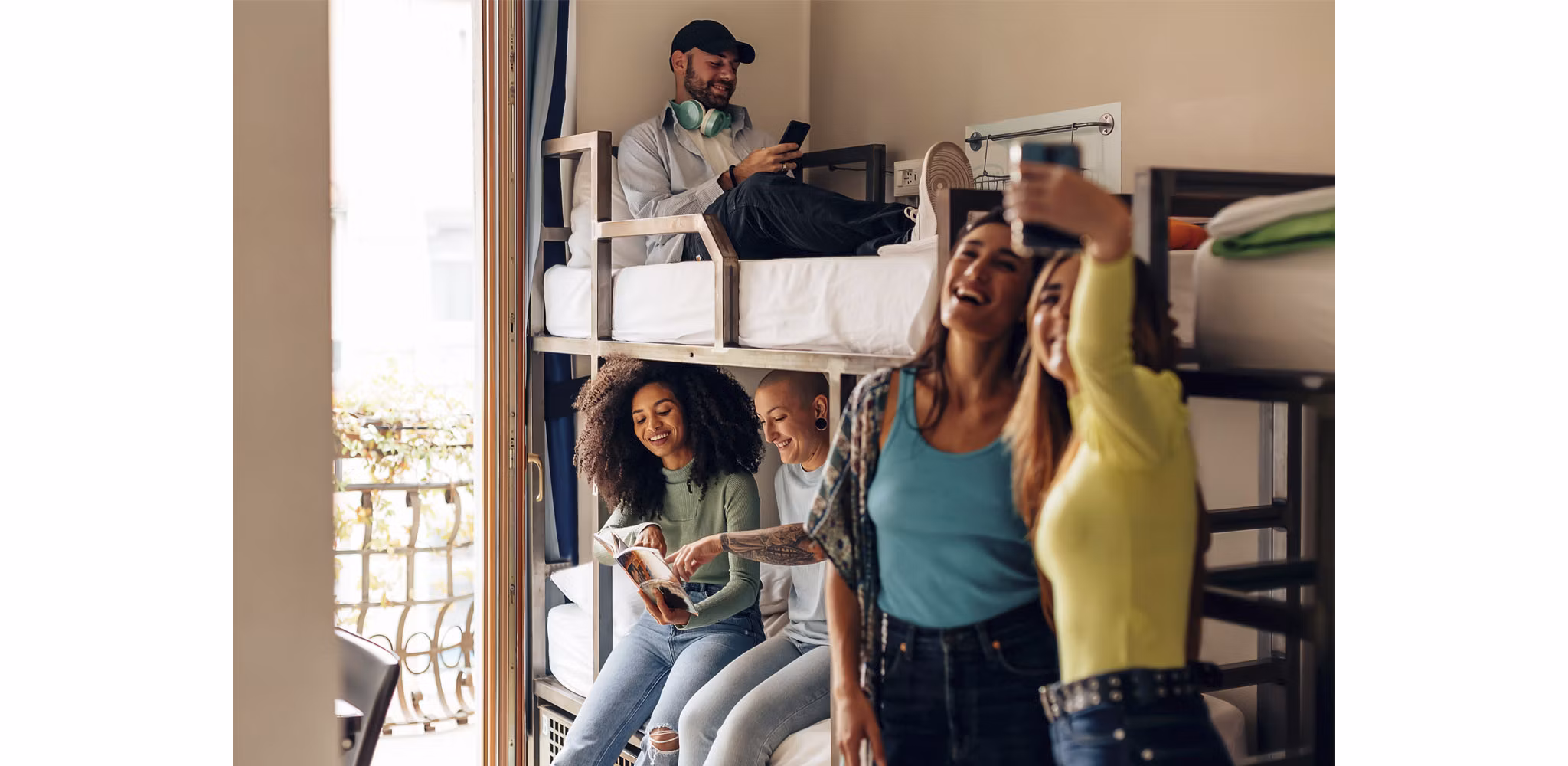 A group of college-aged students in a student housing room with bunk beds, engaging in conversation, using their phones, and reading books.