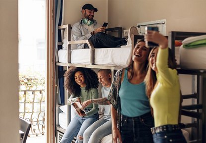 A group of college-aged students in a student housing room with bunk beds, engaging in conversation, using their phones, and reading books.