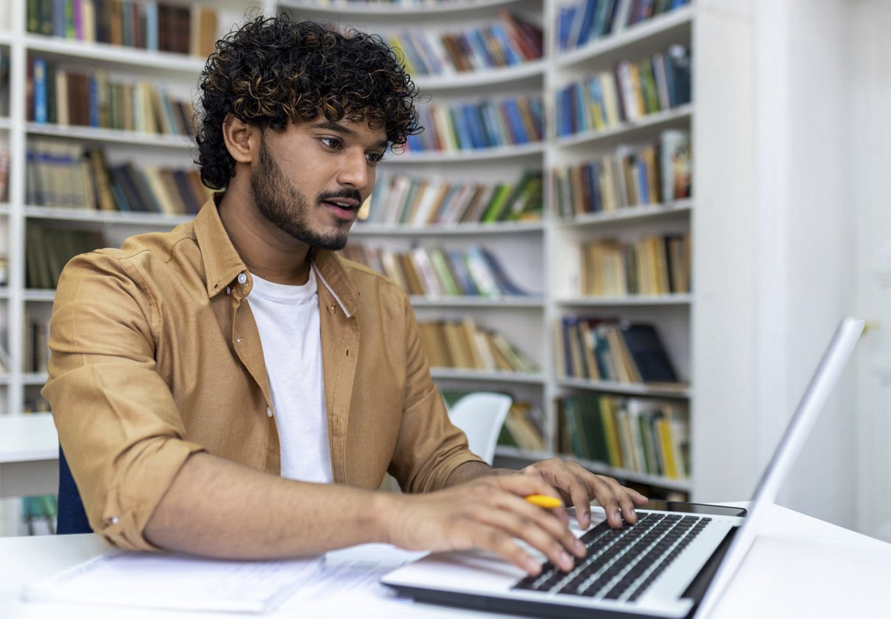 A young man sits at a table in front of a bookcase, typing on his laptop in a modern meeting space within a student housing community.