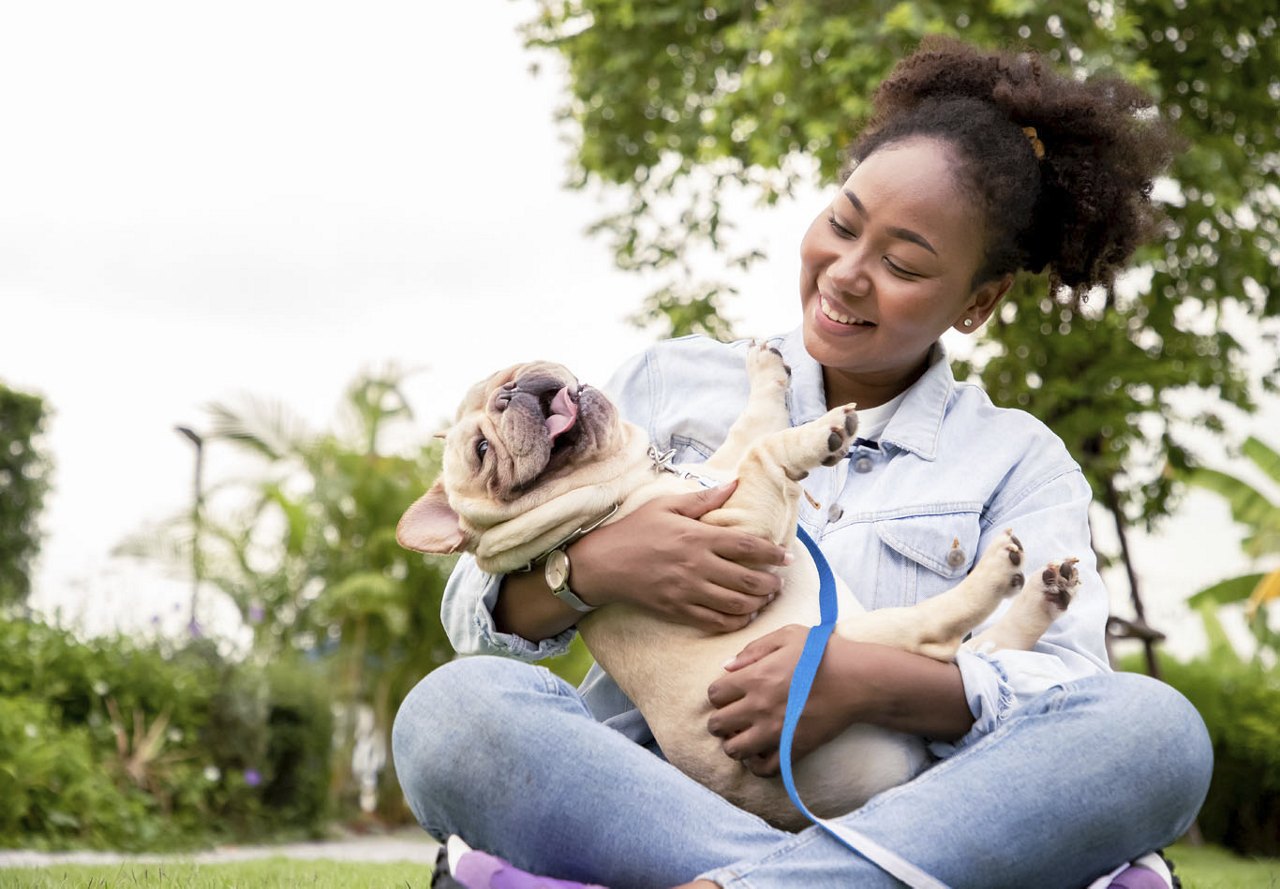 A young African American woman sits on the grass in a park, smiling while holding her French bulldog, with trees and nature in the background.