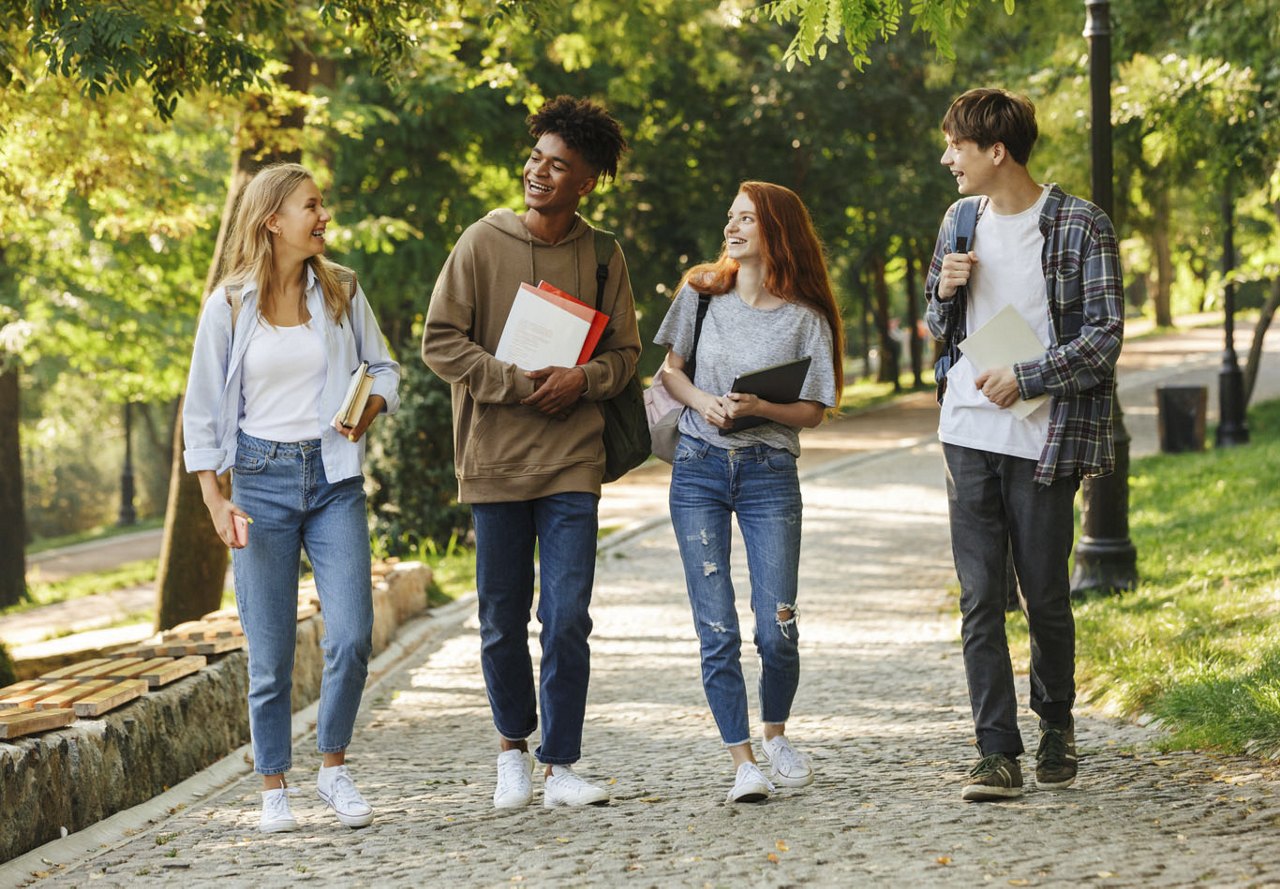 A diverse group of students walks together on campus, laughing and talking as they head to class, surrounded by lush greenery and nature.