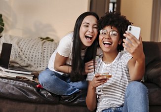 Two roommates smiling as they take a selfie in their cozy living area, enjoying glasses of wine and capturing the joyful moment in their home.