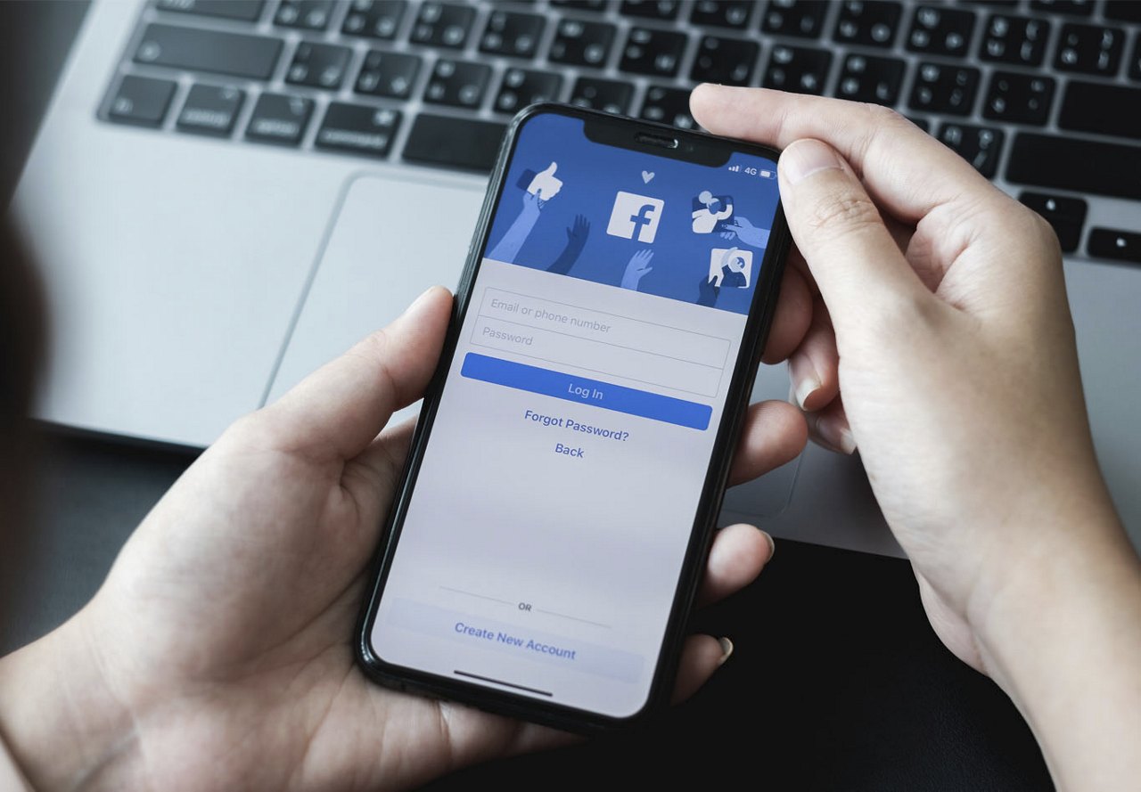  A person looking at their smartphone with the Facebook login screen displayed, while a computer keyboard is visible in the background.