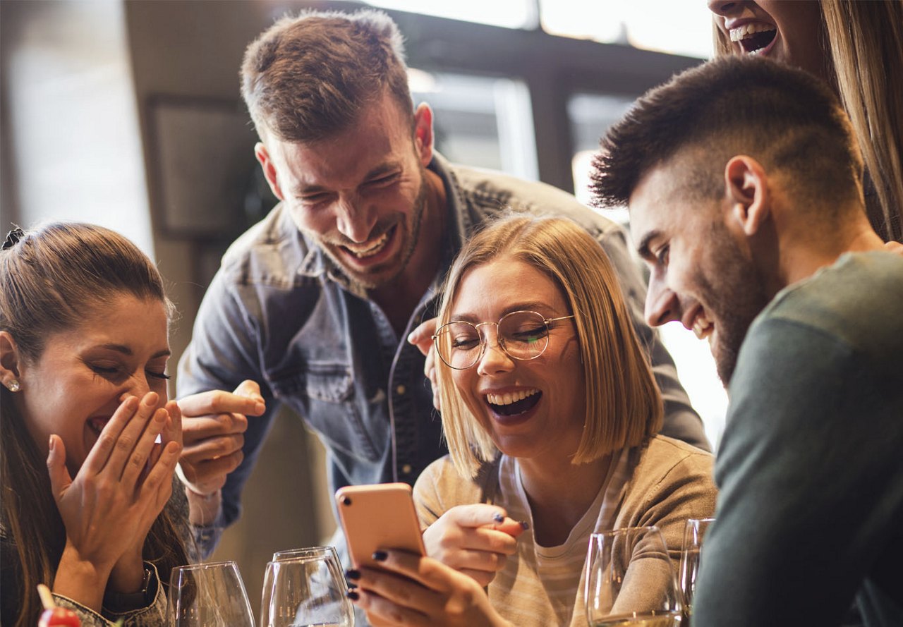 A group of friends smiling and enjoying each other's company, looking at a phone and sipping drinks in a lively restaurant setting.