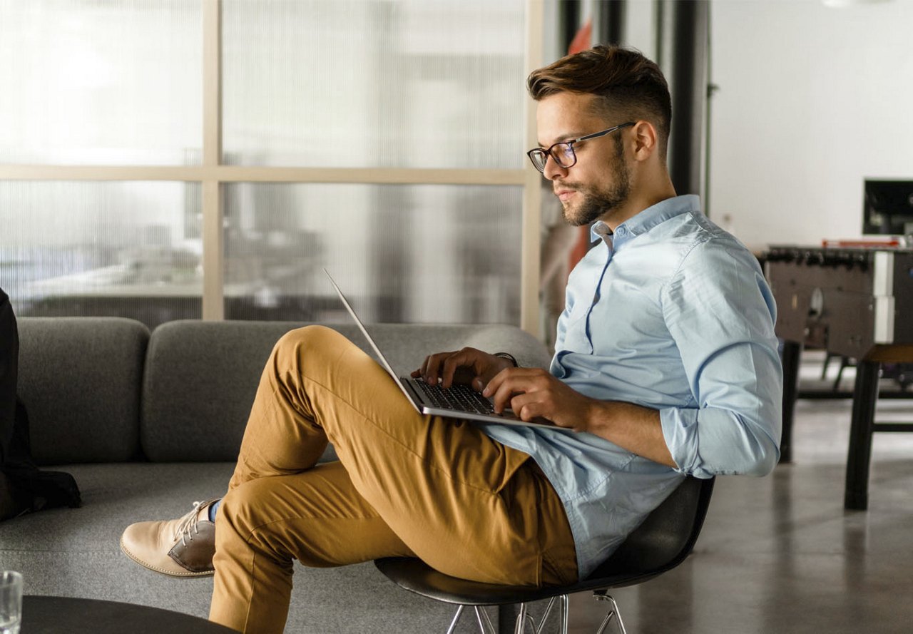 A young man wearing yellow pants and a denim shirt, sitting in his home and focused on researching on his computer.