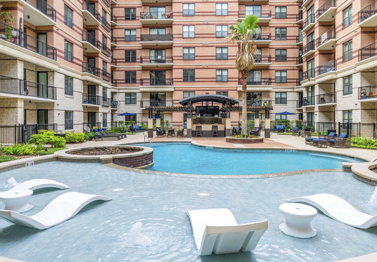 A serene apartment pool in Texas surrounded by plenty of lounge chairs for relaxing, with a beautifully designed building in the background and lush trees and greenery adding to the tranquil atmosphere.