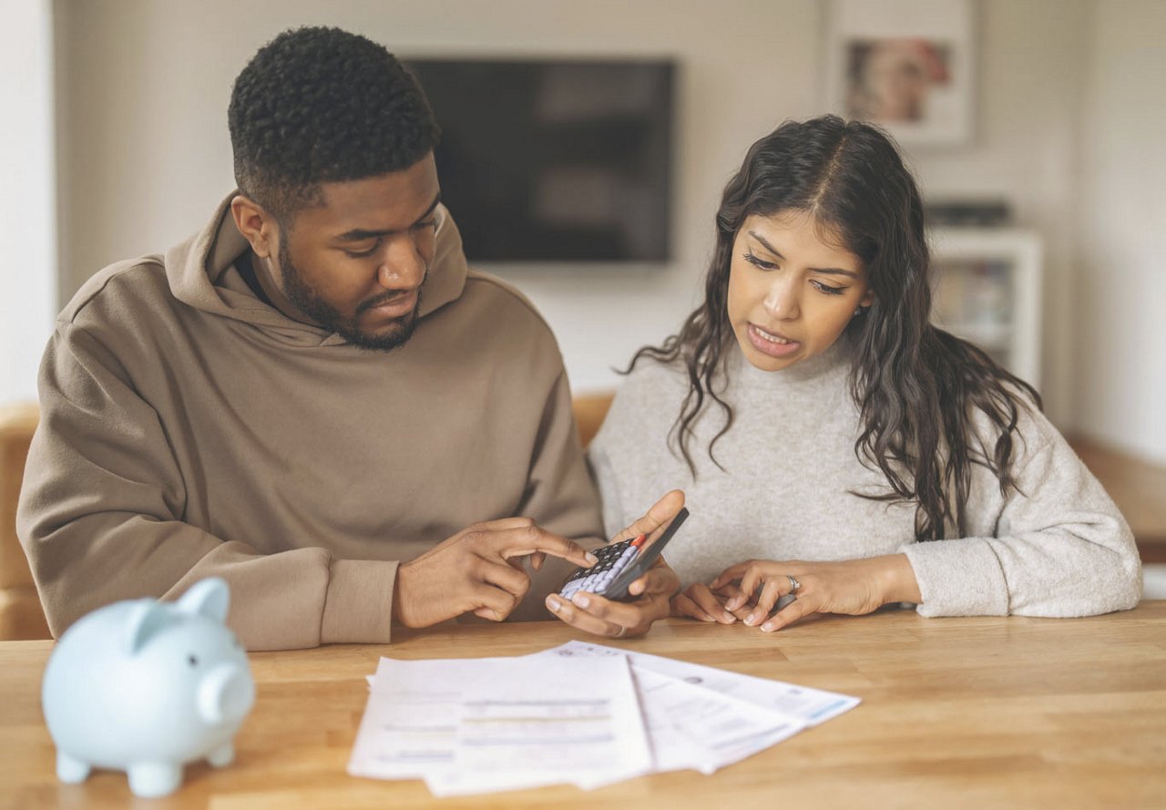 A young couple sitting at a table reviewing their finances, with the man using a calculator as they work together to create a budget.