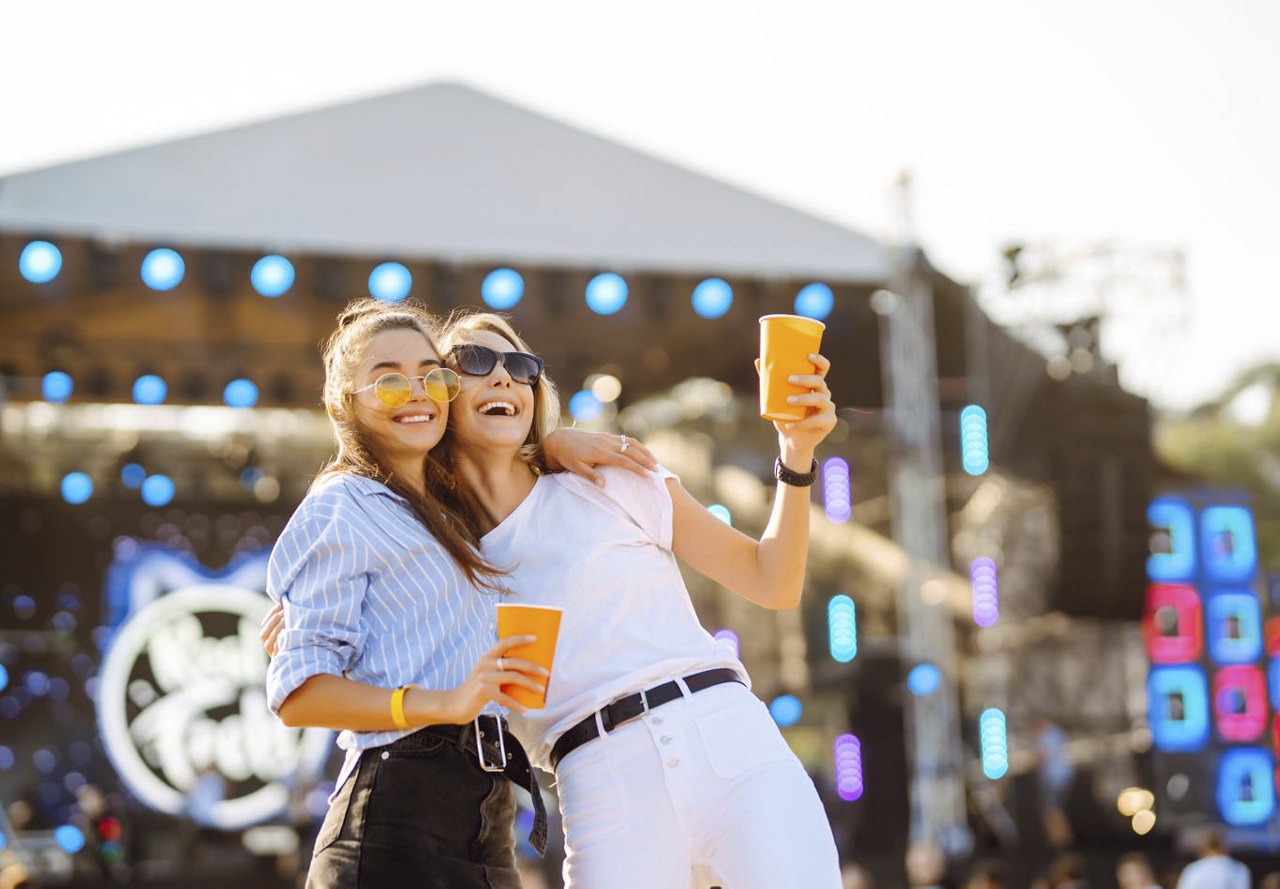 Two female friends enjoying a music festival in Austin, holding drinks and smiling together, with a performance tent featuring artists in the background, creating a lively atmosphere.