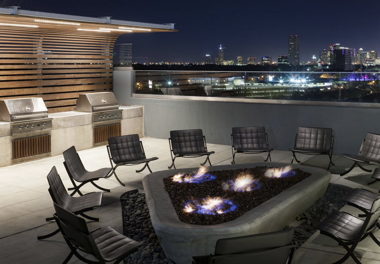 A rooftop area atop an apartment building featuring a cozy fire pit surrounded by comfortable chairs, perfect for outdoor grilling and socializing with friends, all set against a stunning cityscape backdrop.