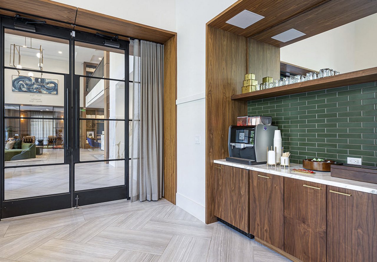 A coffee area in an apartment community featuring modern appliances and warm wooden touches. Through the doors in the background, the lobby is visible, adding to the inviting and stylish atmosphere of the space.