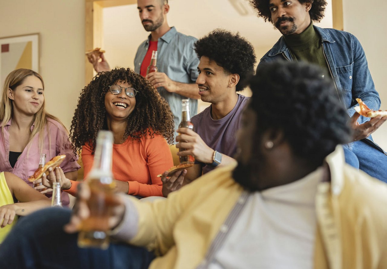 Friends enjoying time together in an apartment community, laughing and sharing drinks and pizza. The lively and relaxed atmosphere highlights their camaraderie and the welcoming environment of their shared space.