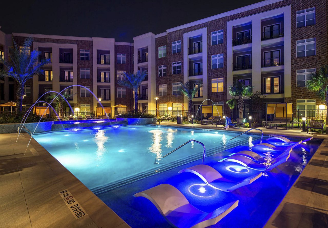 A pool at nighttime at a Greystar Houston apartment complex, illuminated by lights and accented with fountains, with the building's exterior glowing in the background.