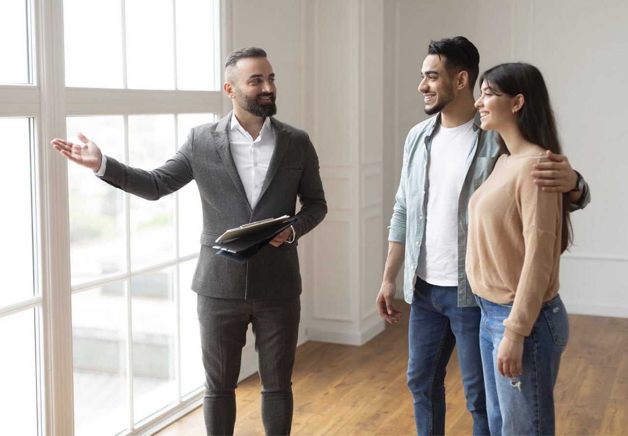 A young couple tours an empty apartment unit with a staff member, standing by the expansive windows to check out the view before deciding on the lease.