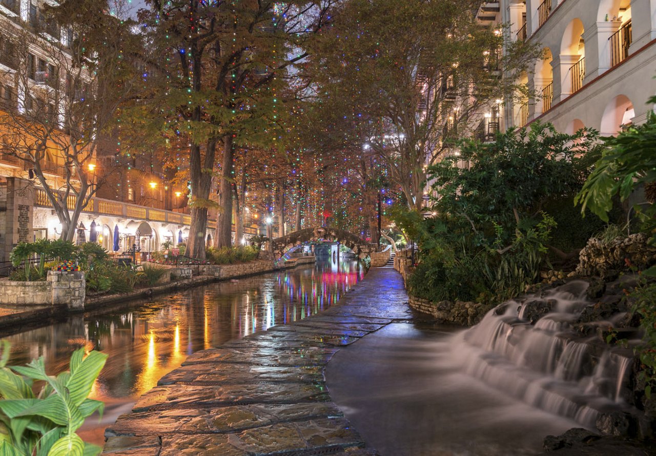 A picturesque evening at the San Antonio River Walk, featuring a canal lined with trees decorated in colorful string lights, while reflections from surrounding buildings shimmer in the water.