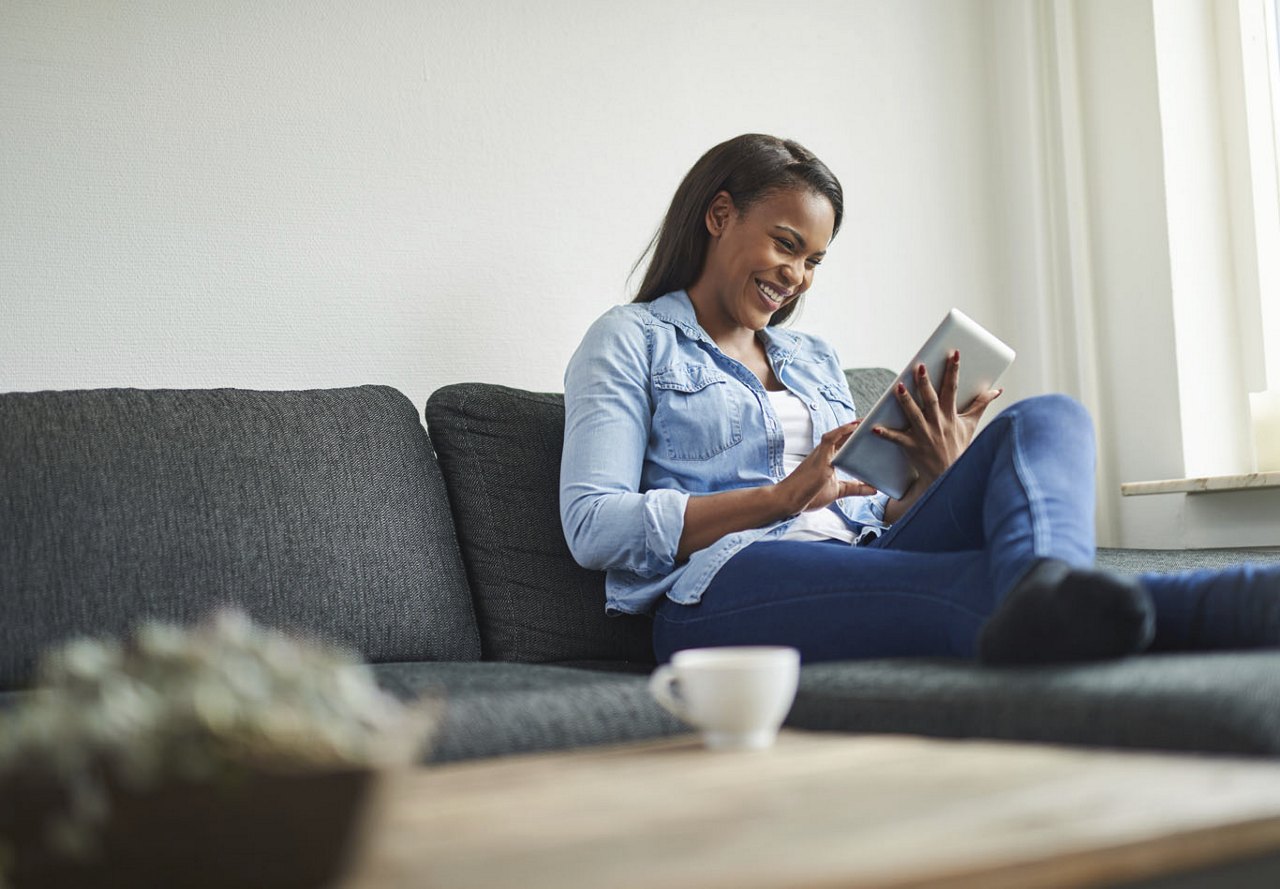 A young African American woman sits on her couch at home, smiling as she looks at her tablet, with blurred decorative items in the foreground.