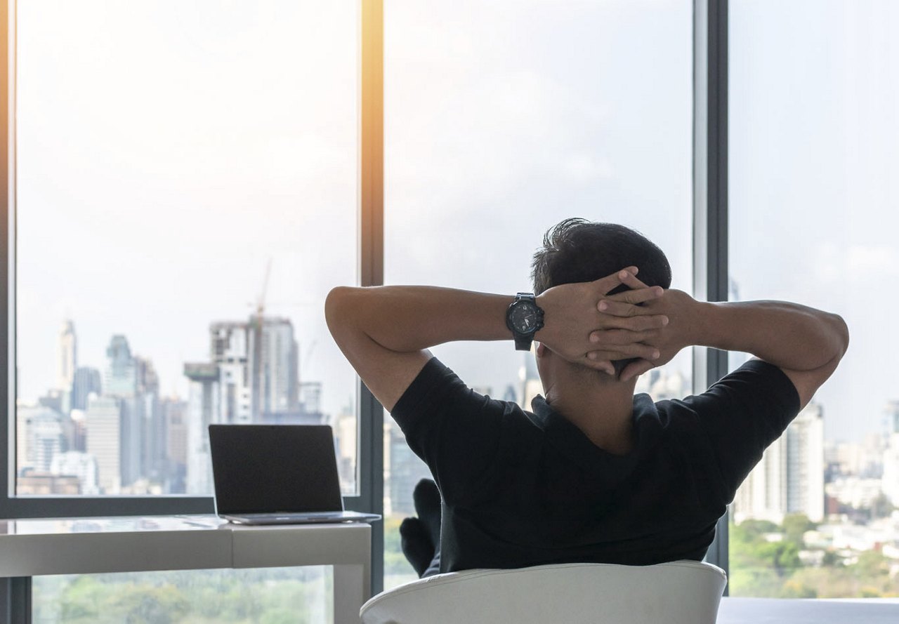 A man sits at his desk in his apartment with his hands folded behind his head, gazing out the large window at the city while working from home.