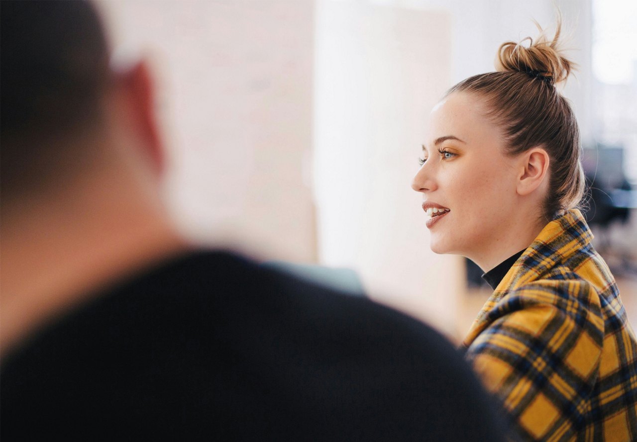 A woman in a bright yellow checkered shirt engaging in conversation with colleagues in an office setting, with another person blurred in the foreground.