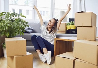 A young woman celebrating with her hands in the air, surrounded by moving boxes, after completing her move. Bright, open windows with flowing curtains provide a sunny backdrop, highlighting her excitement and the fresh start in her new space.