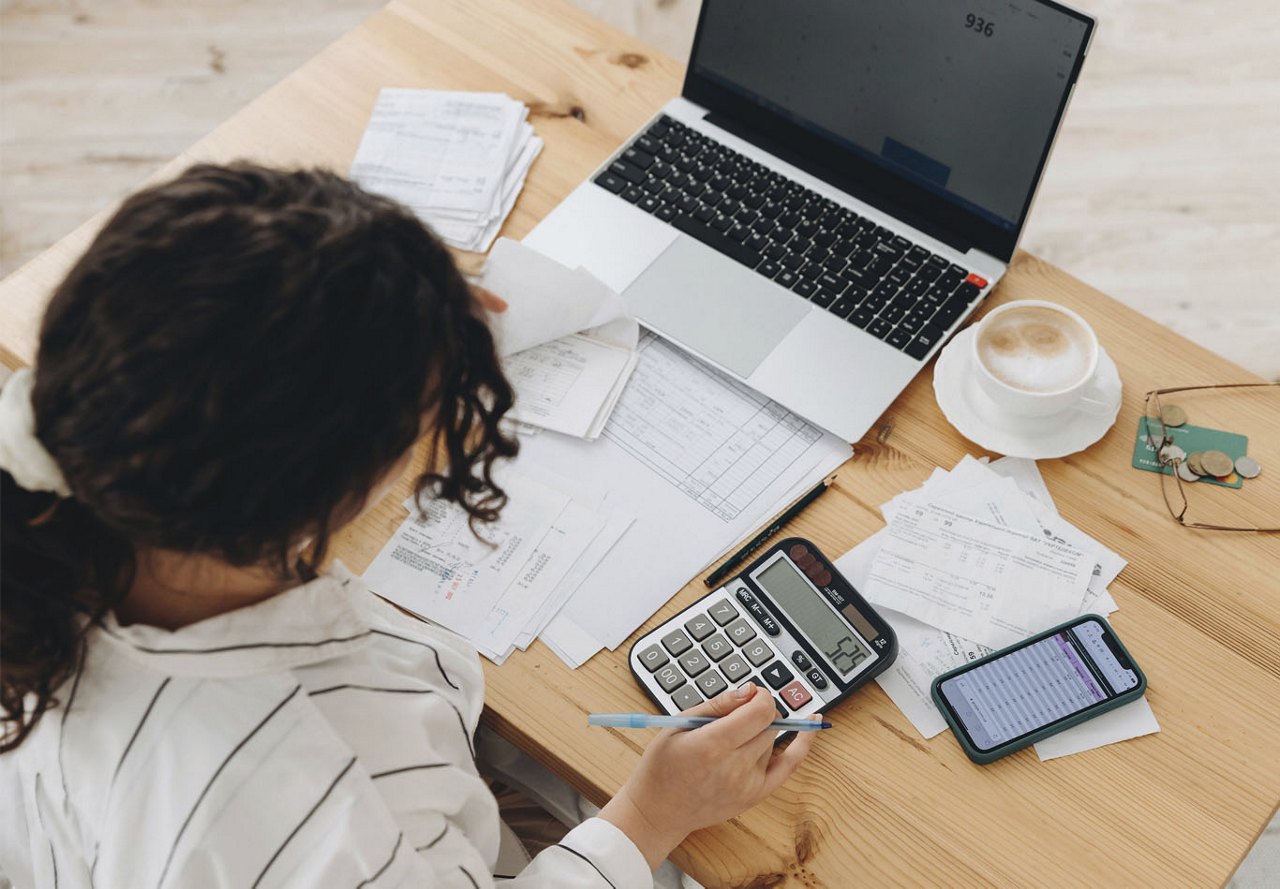 A woman sits at a wooden desk, surrounded by papers, a calculator, and various budgeting tools, actively engaged in financial planning or analysis.