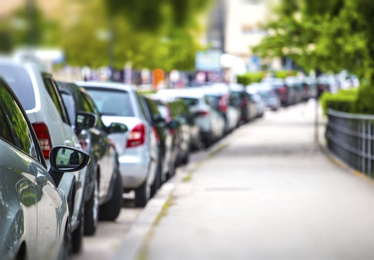 Cars parked in parallel spots along a city street, with a sidewalk next to them and urban buildings in the background.