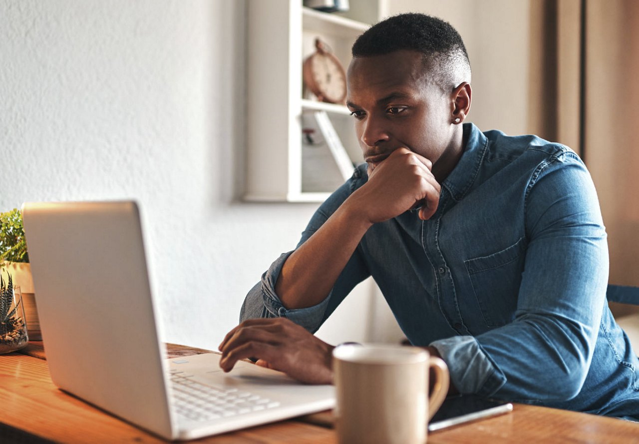 A young African American man sitting at a computer with a cup of coffee, preparing to research his next apartment community. The background features stylish home decor, creating a cozy and focused workspace.