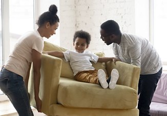 A couple arranging furniture in their new home while their young son sits happily in the yellow armchair they’re carrying, smiling as they settle into the space together.