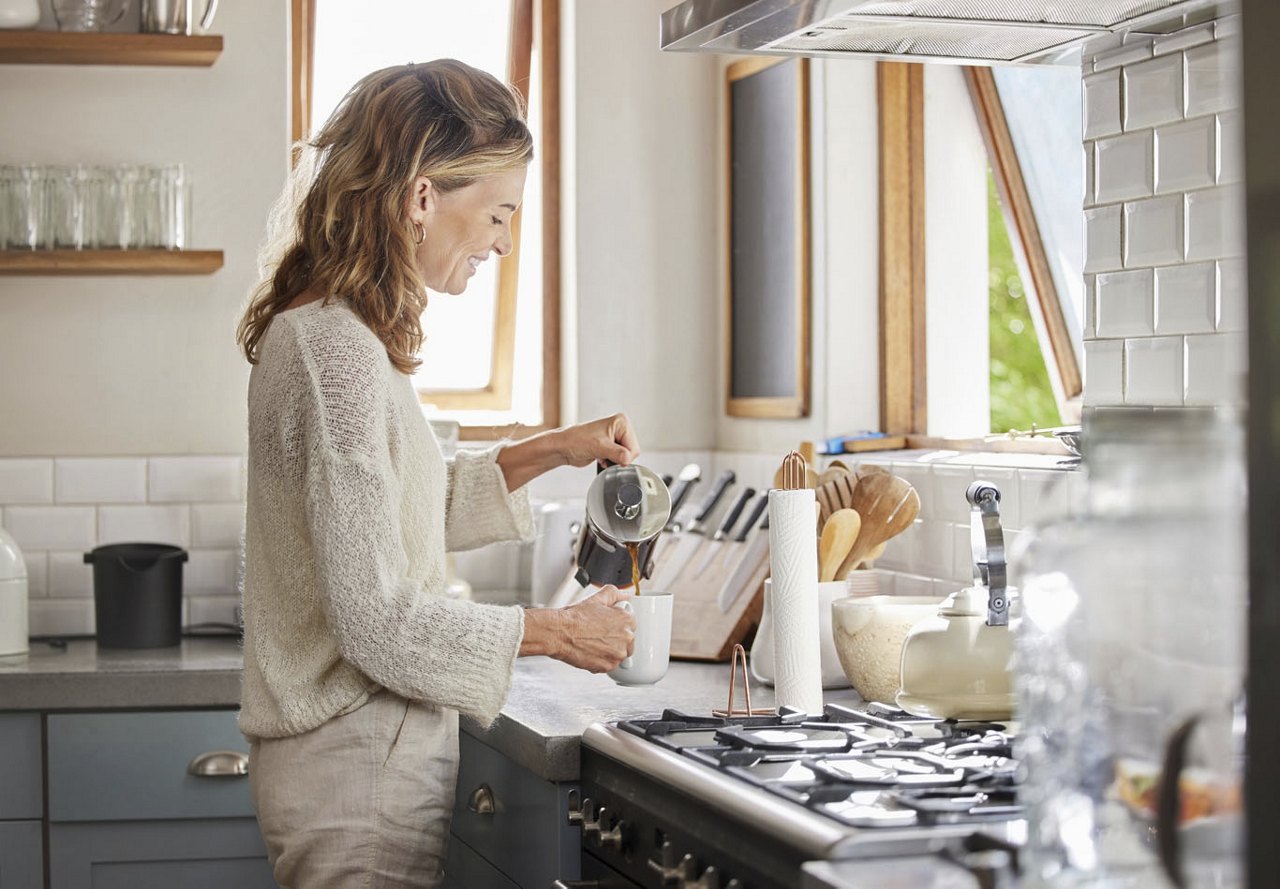 An older woman in a kitchen filled with warm decor and modern appliances, pouring coffee into a mug as she begins her day. 