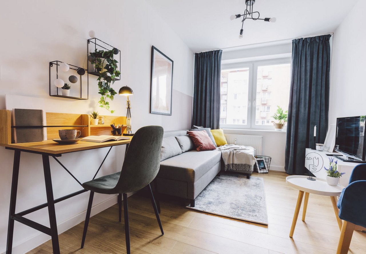 A modern living area filled with natural light from a large window, featuring a cozy workspace with a desk. The room is decorated with curtains, a patterned rug, and accent pillows, creating a bright and inviting atmosphere.