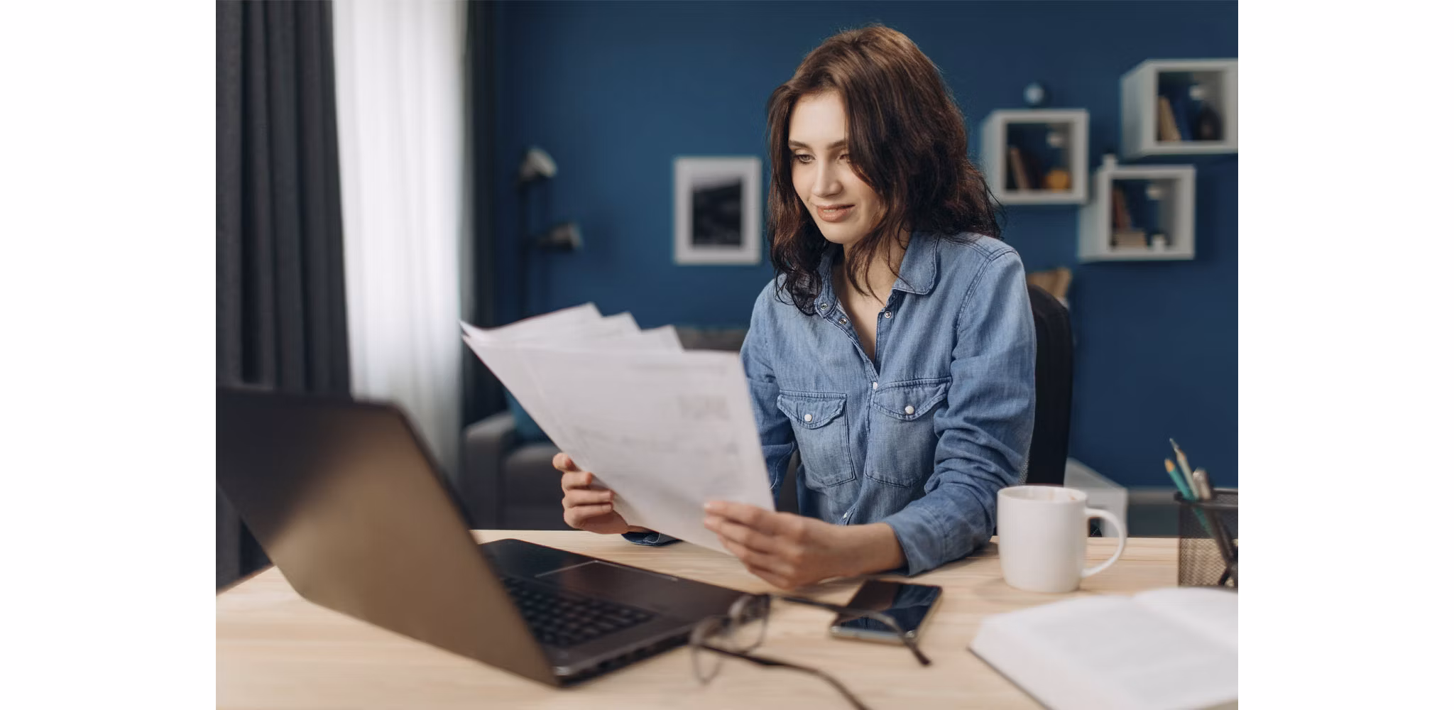 A woman reviews papers at her desk, with a computer, glasses, a phone, and a coffee mug nearby. Behind her is a blue wall adorned with various decorations.