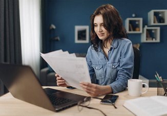 A woman reviews papers at her desk, with a computer, glasses, a phone, and a coffee mug nearby. Behind her is a blue wall adorned with various decorations.