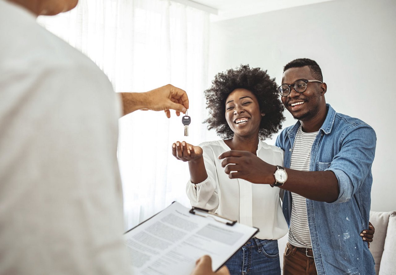 An African American couple holding out their hands to receive the keys to their new apartment from the community manager. Both are smiling with excitement, celebrating the start of their new journey.