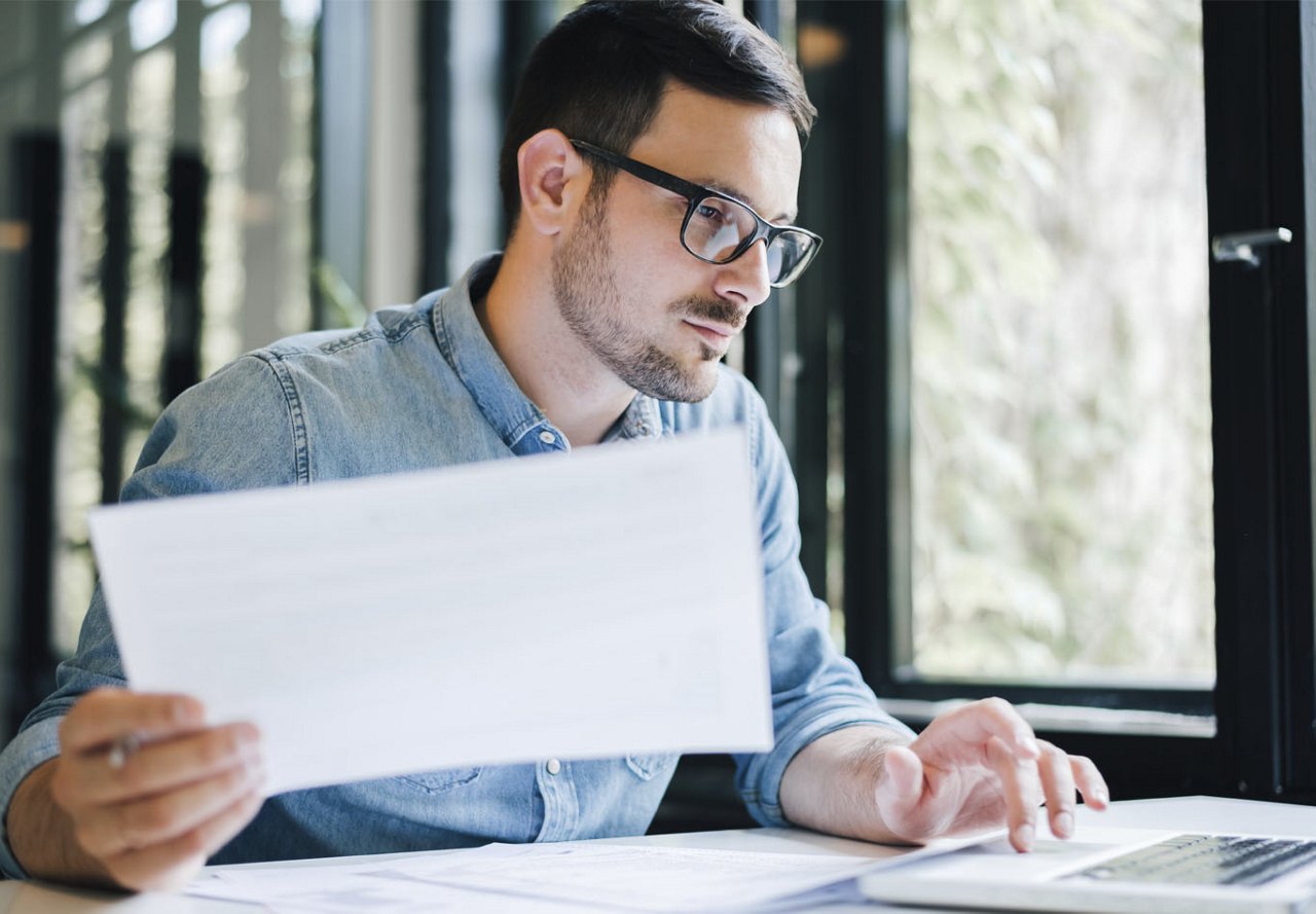 A man sitting at a desk, organizing paperwork and looking at his computer as he prepares to submit a rental application. Open windows in the background let in natural light, creating a bright and focused workspace.