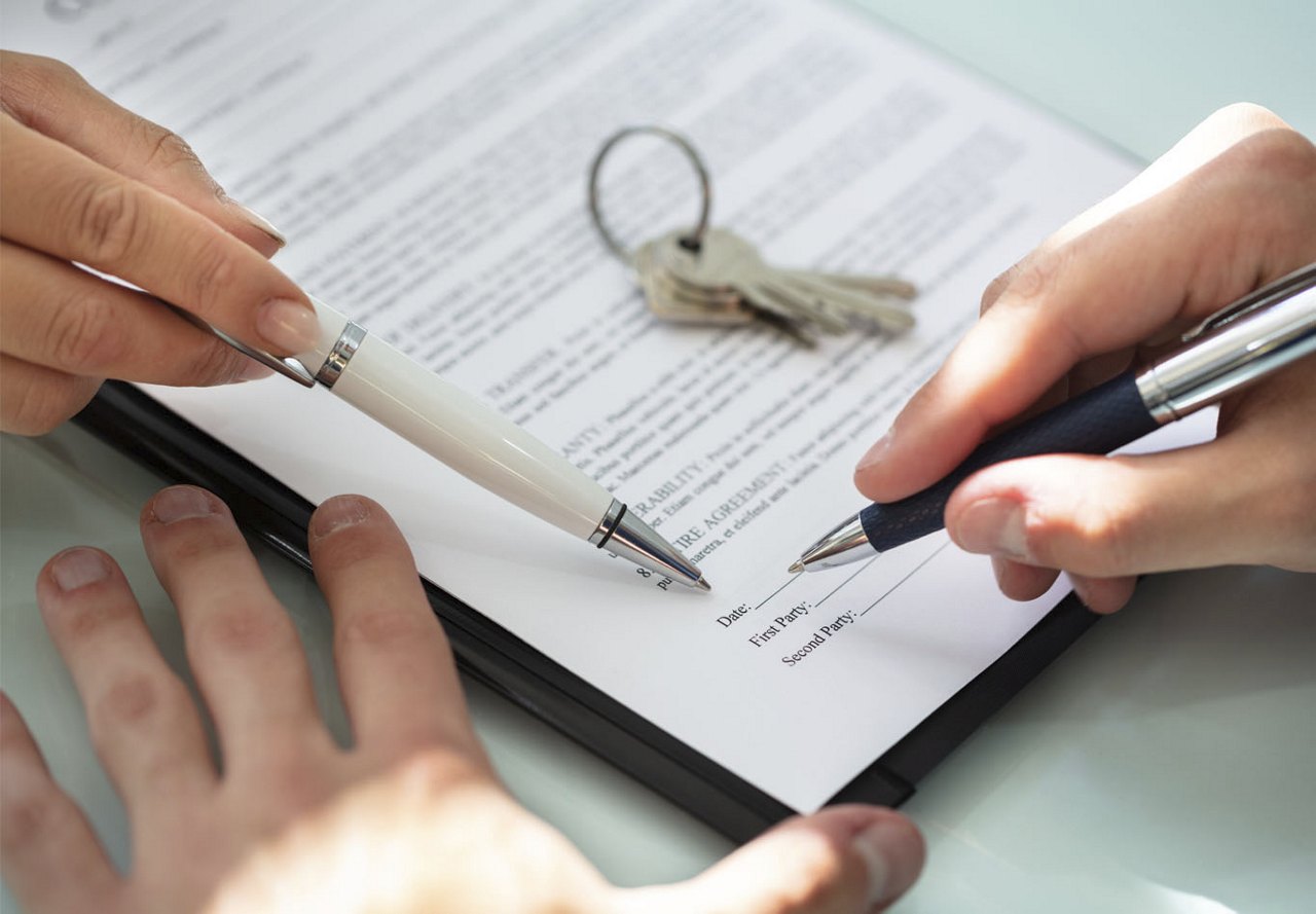 Hands pointing to a line on a rental agreement as someone signs it, with keys placed on the paper ready to be handed over once the agreement is complete.