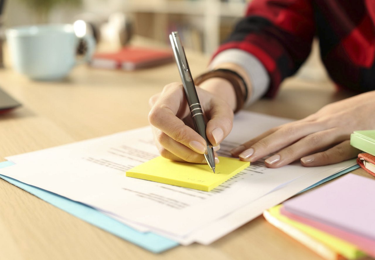 A person writing on a sticky note with a pen at a desk, surrounded by paperwork underneath and various desk items in the background, creating an organized and focused workspace.