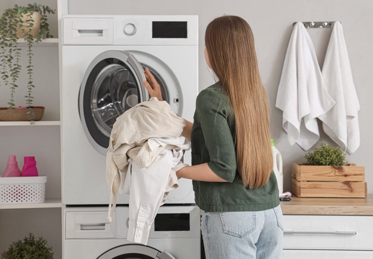 Woman facing the laundry machines, loading clothes to be washed, with cleaning supplies like detergent and towels around her in the laundry area of her home.