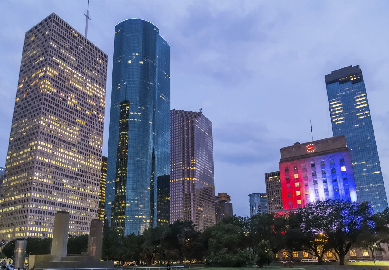 Skyscrapers in the downtown Houston skyline at night, featuring a state building illuminated with red, white, and blue lights, and lights filling all the windows of the building as the sky darkens.