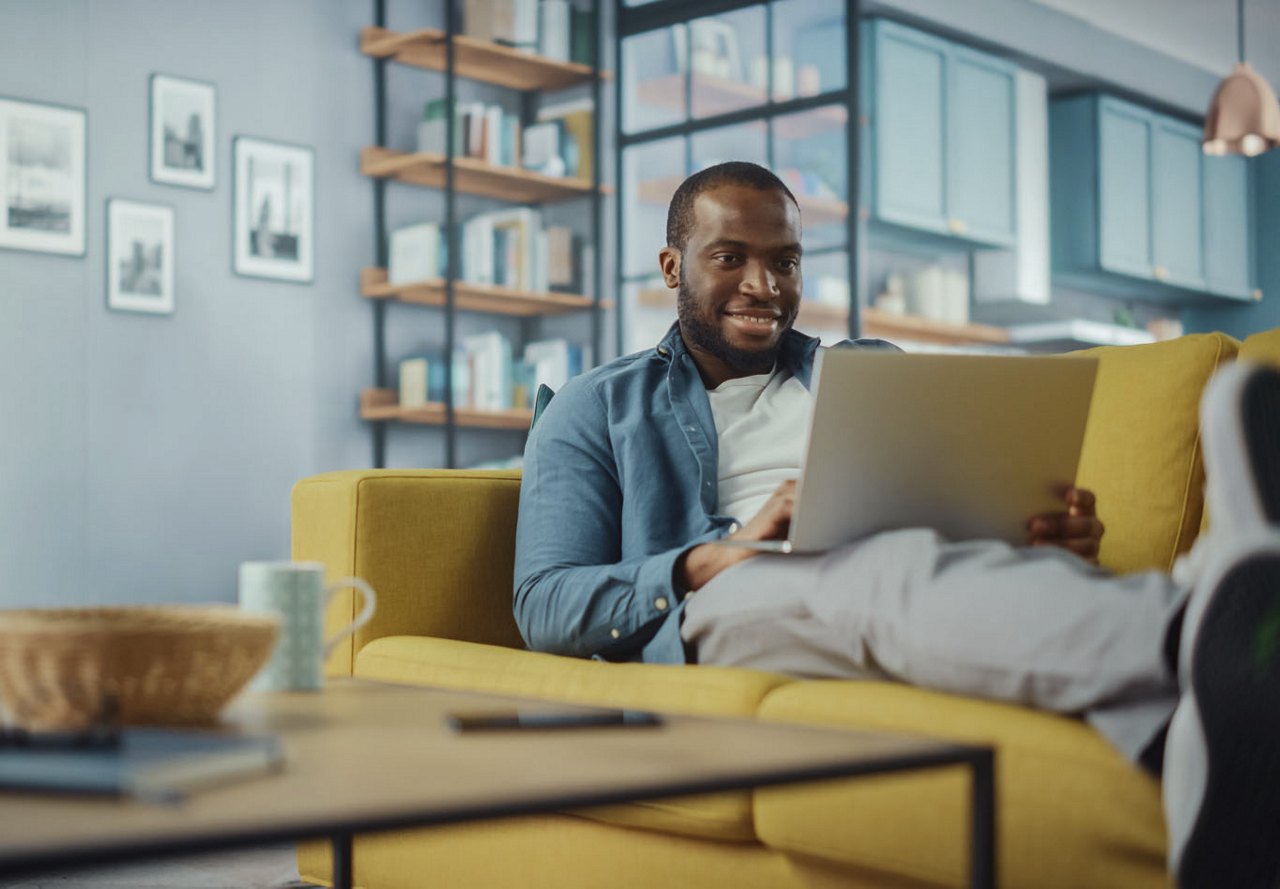 African American man sitting on a yellow couch in his home, smiling as he looks at his computer screen, surrounded by various home decor items in the room and background.