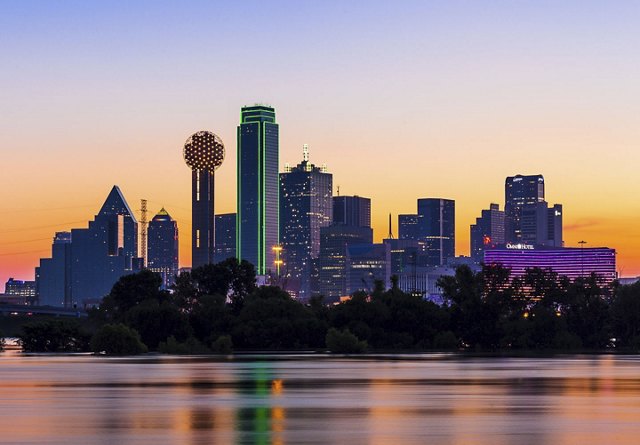 A nighttime view of Dallas, showcasing the city skyline illuminated by neon lights, set against a colorful sunset backdrop. The water in the foreground reflects the vibrant city lights, adding to the dramatic and lively scene.