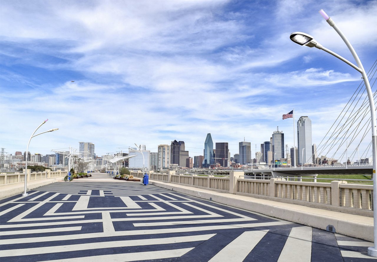  A bridge in Dallas overlooking the city skyline, framed by a clear blue sky with scattered clouds. The road markings are visible on the bridge, guiding the path toward the vibrant urban backdrop.