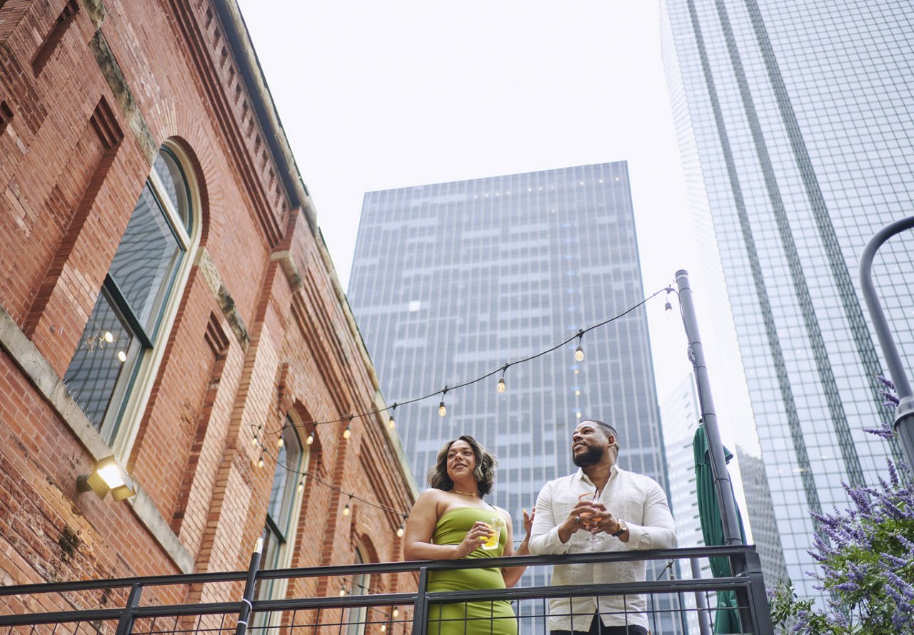 A couple standing together on a balcony, enjoying drinks and taking in the city view below. The balcony is adorned with café lighting hanging from the brick building above, adding a warm and inviting ambiance to their experience.