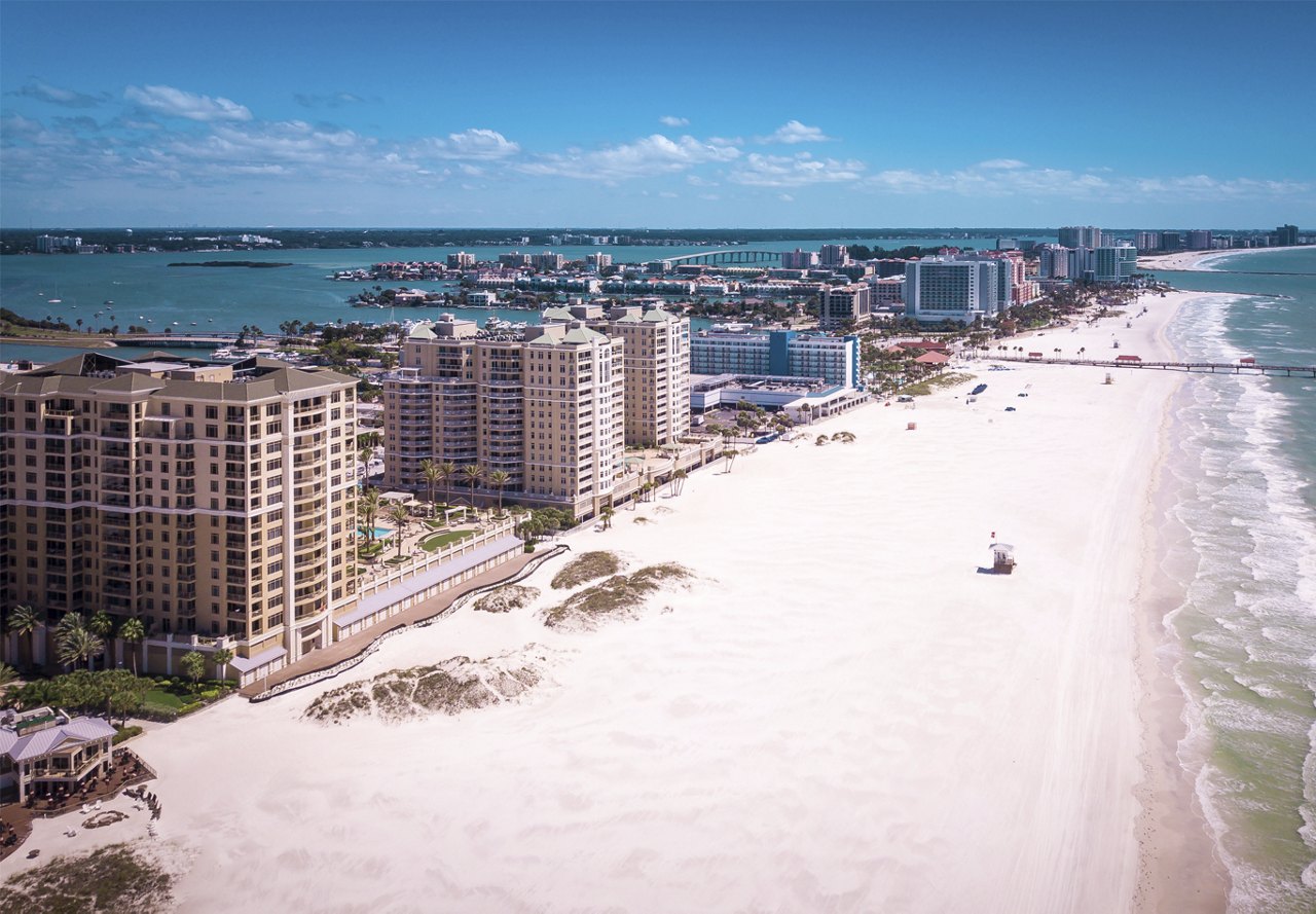 Beach and buildings along the coastline in Clearwater, Florida, with very few people, surrounded by turquoise water and a clear sky.