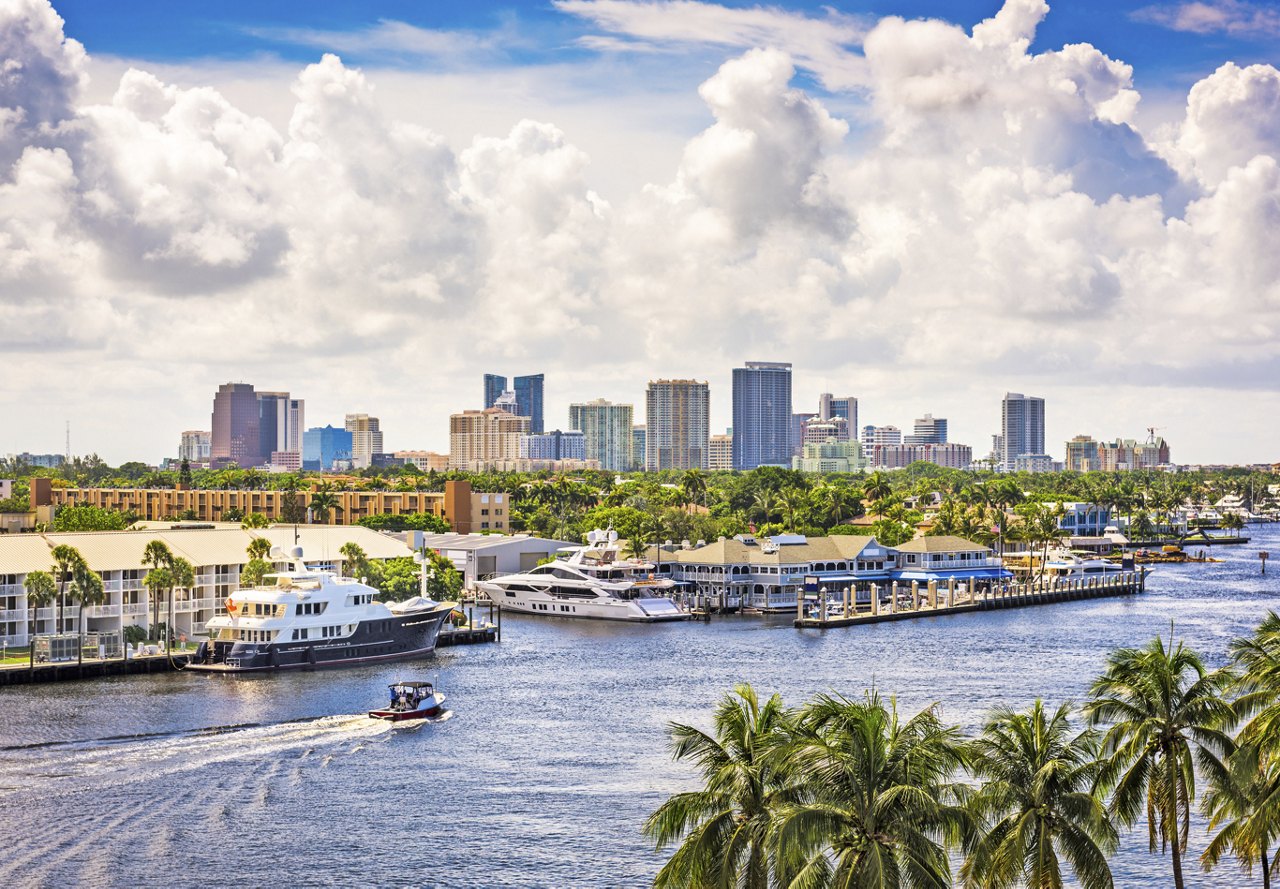 View of Fort Lauderdale's cityscape with water and docked boats, palm trees, and a clear, blue sky.