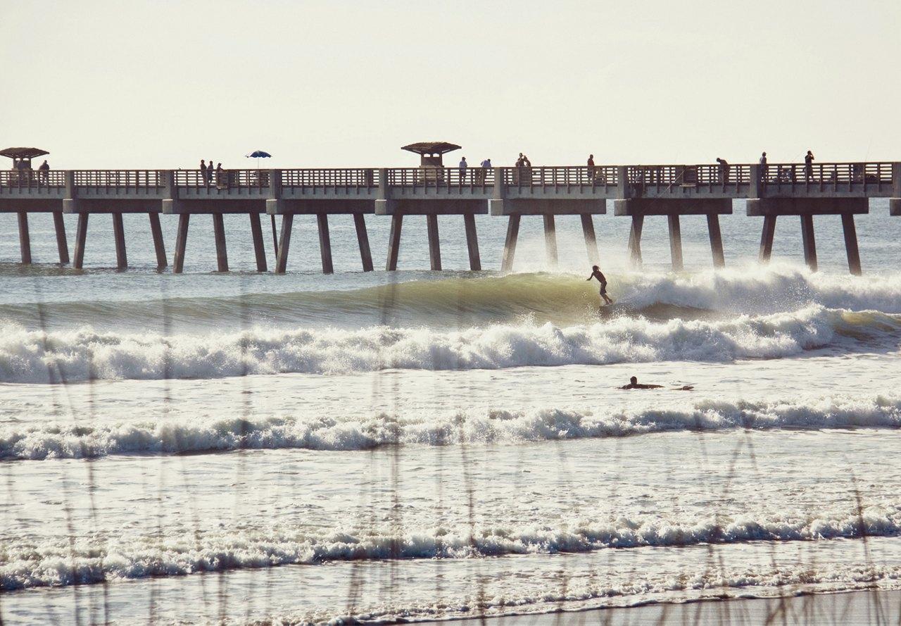 Beach view in Jacksonville, Florida, featuring a surfer riding the waves and a pier with people strolling up and down.