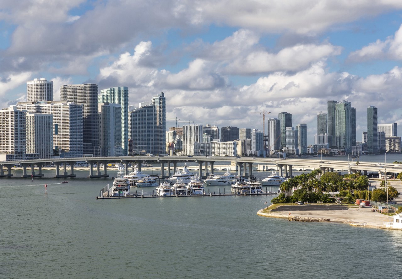 View of Miami in the distance, with a marina filled with boats in the foreground, a bridge connecting to the city, and a backdrop of clouds and a clear sky.