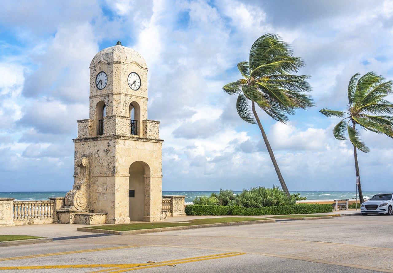 View of a clocktower in Palm Beach, Florida, with palm trees swaying in the wind, a clear sky in the background, and a street in front of the tower with one car parked to the side.