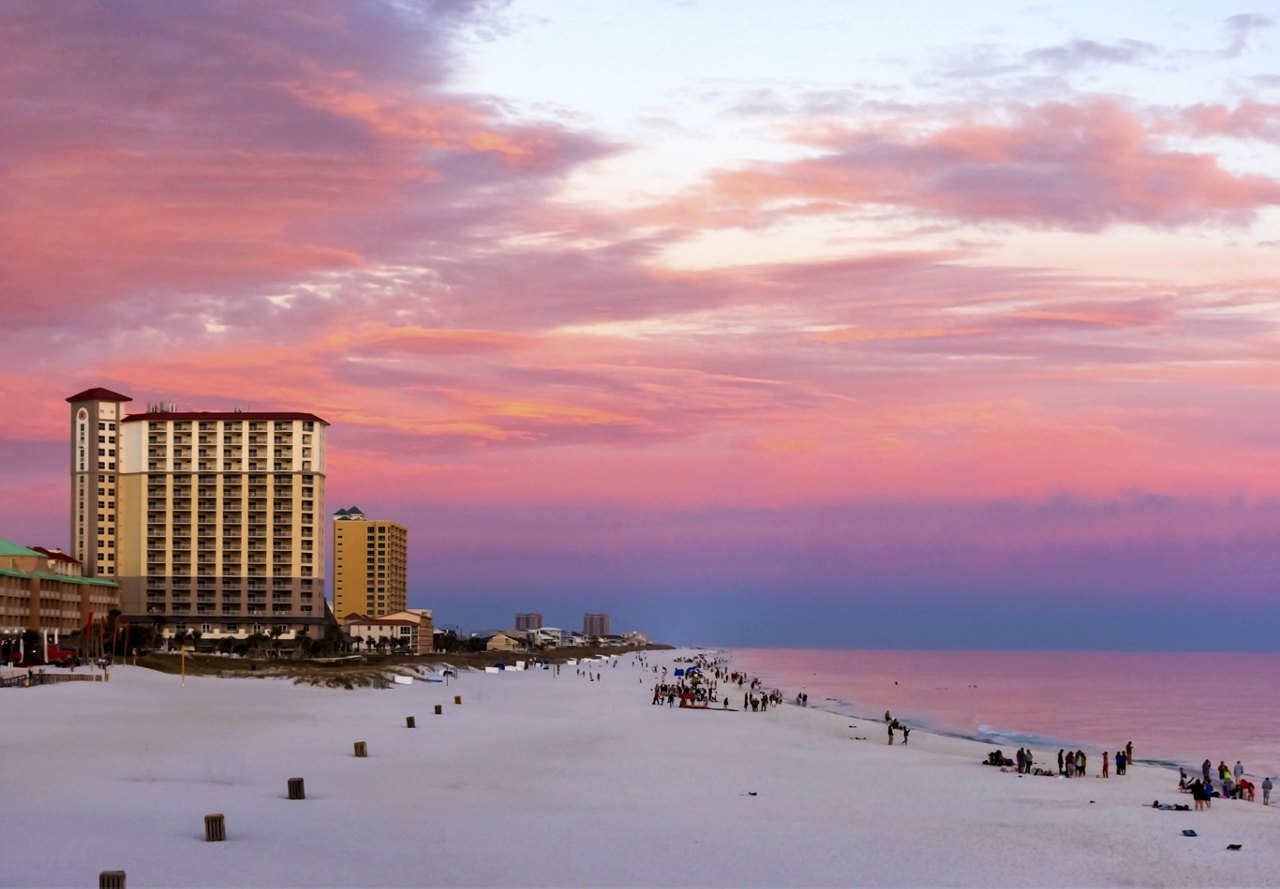 Gorgeous pink and purple sunset on the beach in Pensacola, Florida, with groups of people scattered across the sand and large structures visible behind the beach.