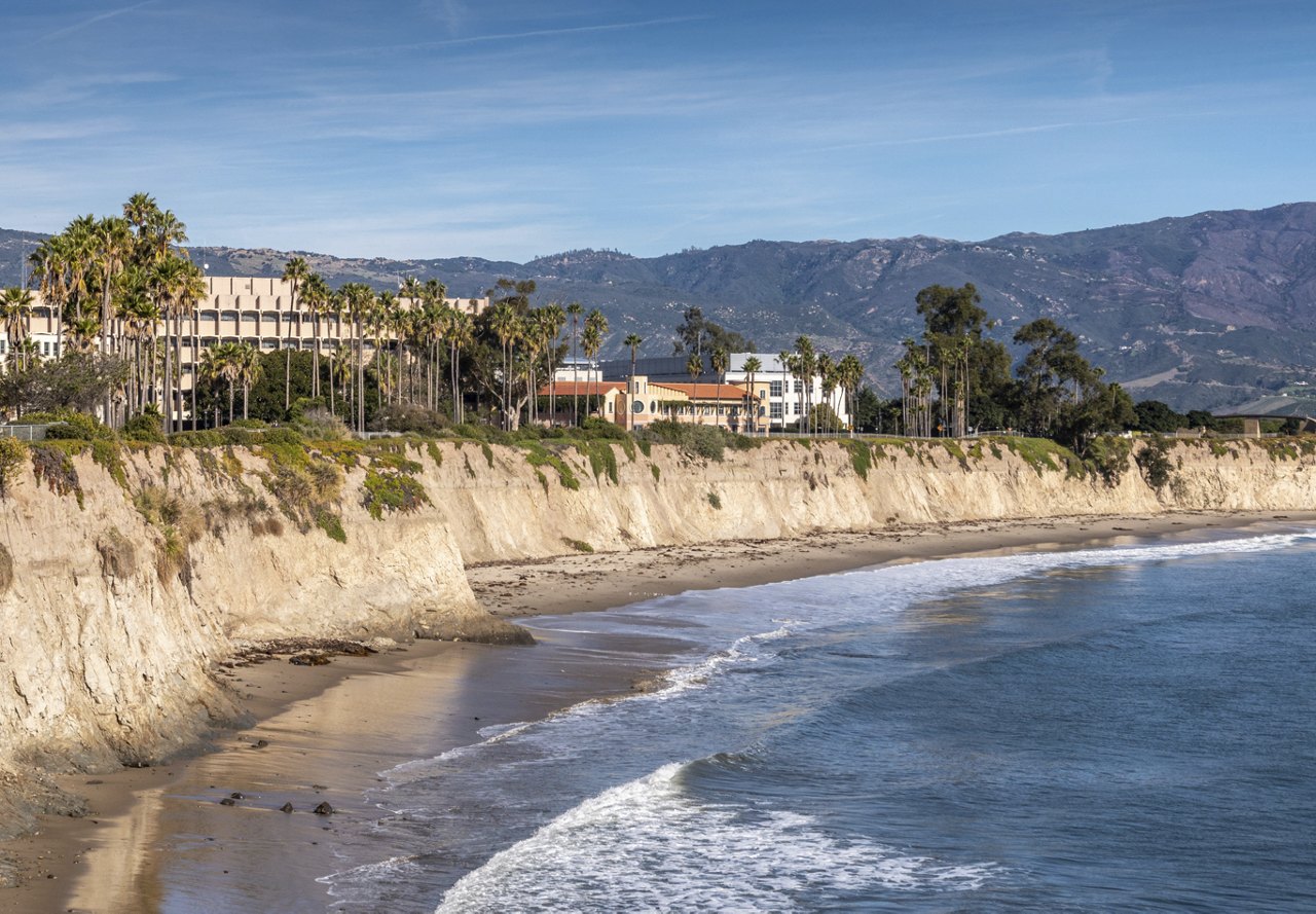 A scenic view of the Santa Barbara coastline, featuring houses along the shore with mountains in the background and the water gently lapping at the beach.